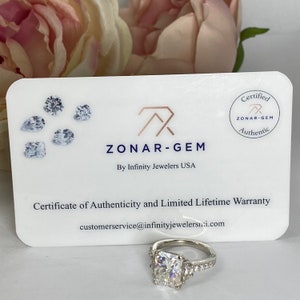 May include: A white card with the text "ZONAR-GEM" and "By Infinity Jewelers USA" and a certificate of authenticity and limited lifetime warranty. The card also has a round seal with the text "ZONAR-GEM" and "Authentic". A silver ring with a large square-shaped diamond is in the foreground.