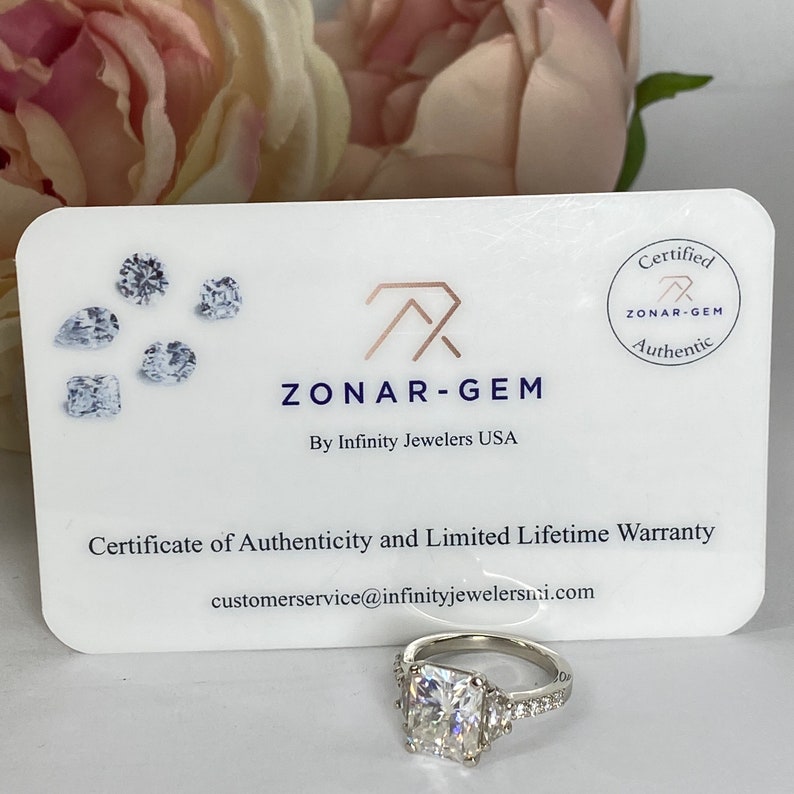 May include: A white card with the text "ZONAR-GEM" and "By Infinity Jewelers USA" and a certificate of authenticity and limited lifetime warranty. The card also has a round seal with the text "ZONAR-GEM" and "Authentic". A silver ring with a large square-shaped diamond is in the foreground.