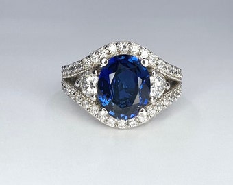 Op de afbeelding: Een zilveren ring met een grote blauwe saffier omringd door een halo van kleinere diamanten. De ring heeft een dubbele band met extra diamanten aan de zijkanten.
