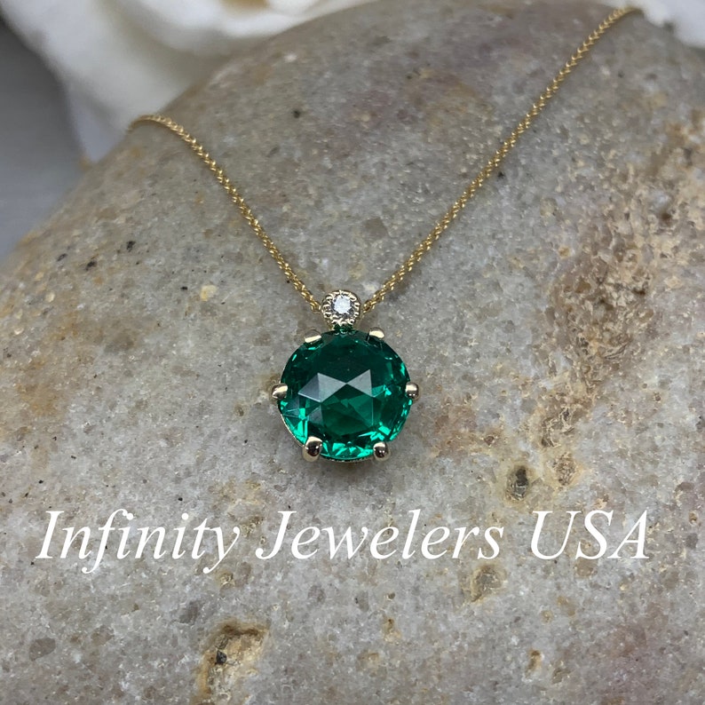 May include: A gold chain necklace with a green gemstone pendant. The pendant is a round, faceted stone with a smaller diamond above it. The necklace is on a gray, textured surface. Infinity Jewelers USA is written below the pendant.