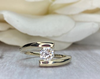 May include: A gold engagement ring with a single round diamond set in a unique, modern design. The band is thin and sleek, with a curved, open-ended design that allows the diamond to sit prominently.