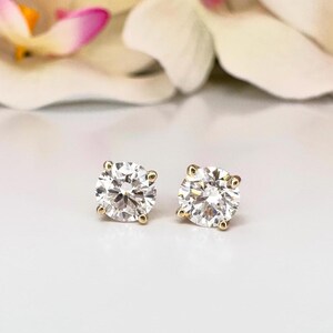 May include: A pair of round-cut diamond stud earrings with gold settings. The diamonds sparkle brightly against a soft, white background, with a blurred floral element in the background. The earrings are a classic jewelry item.
