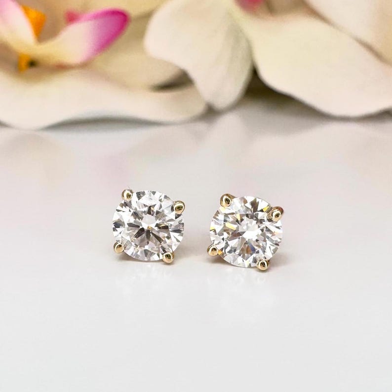May include: A pair of round-cut diamond stud earrings with gold settings. The diamonds sparkle brightly against a soft, white background, with a blurred floral element in the background. The earrings are a classic jewelry item.