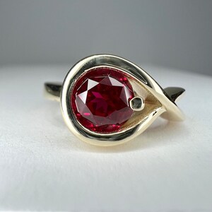 Puede incluir: Un anillo de oro con una gran gema roja oscura facetada. El anillo tiene un diseño único y fluido con una piedra de acento gris más pequeña. El engaste es de color oro cálido, creando un contraste llamativo con la gema roja.