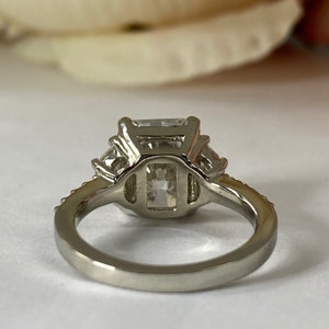 May include: A silver ring with a large, square-shaped, clear gemstone surrounded by smaller, clear gemstones. The ring has a simple band.