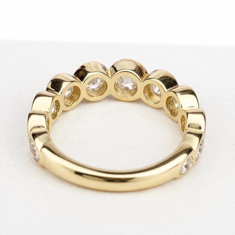 Puede incluir: Un anillo de oro con una serie de piedras preciosas redondas y transparentes engastadas en la parte superior. El anillo tiene un acabado pulido y las piedras est&aacute;n espaciadas uniformemente. El anillo se muestra sobre un fondo blanco.