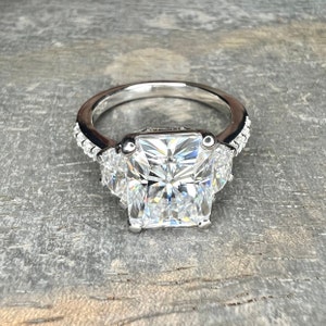 May include: A silver ring with a large square-shaped diamond in the center and two smaller triangular diamonds on either side. The ring is set with smaller diamonds around the band.