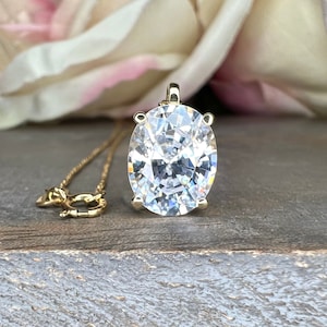 May include: A gold pendant necklace featuring a large, oval-cut, clear gemstone. The pendant is suspended from a delicate gold chain. A small gold dachshund charm is also on the chain. The background includes a blurred flower and a wooden surface.
