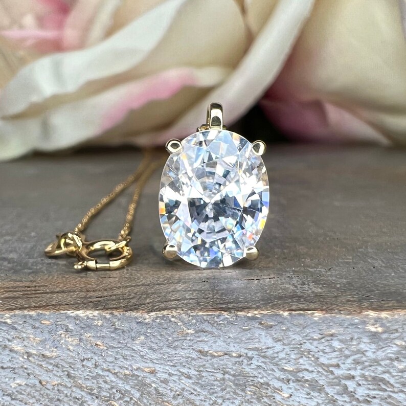 May include: A gold pendant necklace featuring a large, oval-cut, clear gemstone. The pendant is suspended from a delicate gold chain. A small gold dachshund charm is also on the chain. The background includes a blurred flower and a wooden surface.