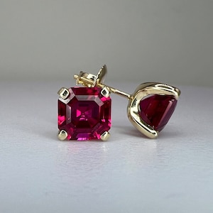 May include: A pair of gold earrings with square-shaped ruby gemstones. The earrings have a post and push-back closure.