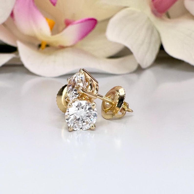 May include: A pair of gold stud earrings with a large, round, clear gemstone as the focal point. Each earring has a smaller cluster of clear gemstones above the main stone. The earrings are set against a soft, blurred background with white and pink floral accents.
