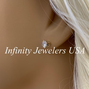 May include: A gold-colored stud earring with a clear, teardrop-shaped gemstone. The earring is set in a gold metal frame and is worn on a person's ear. The text "Infinity Jewelers USA" is visible.