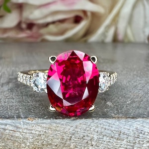 May include: A ring with a large oval-shaped ruby gemstone surrounded by a halo of smaller diamonds. The ring is set in a white metal band.