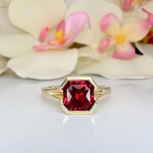 May include: A gold ring with a square-shaped, deep red gemstone set in a gold bezel setting.