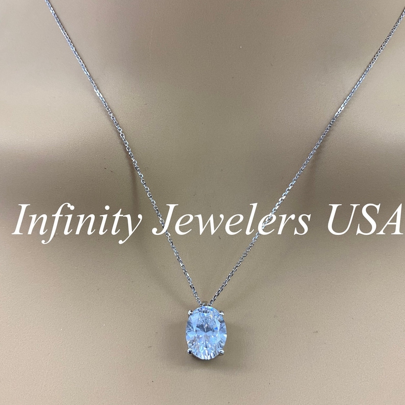 May include: A silver necklace with a large, oval-cut, clear gemstone pendant. The chain is delicate, and the pendant is set in a silver setting. The text "Infinity Jewelers USA" is displayed in white.