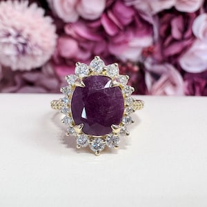 May include: A gold ring featuring a large, oval-cut, deep red gemstone surrounded by a halo of smaller, clear gemstones. The ring's band has a textured design. The background is blurred, with pink and purple flowers.