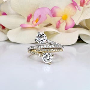 May include: Three diamond rings with a floral background. The rings are silver and gold, with a large, round diamond as the focal point. The bands are encrusted with smaller diamonds. The background features white and pink flowers.