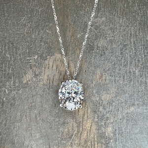May include: A silver necklace with a large, oval-cut, clear gemstone pendant. The pendant is the focal point, reflecting light and set in a four-prong setting. The chain is delicate and hangs against a gray wooden background.