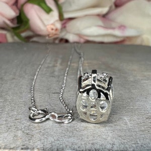 May include: A silver necklace with a pendant featuring a geometric design with clear, sparkling stones. The necklace is displayed on a gray surface with a blurred floral background.