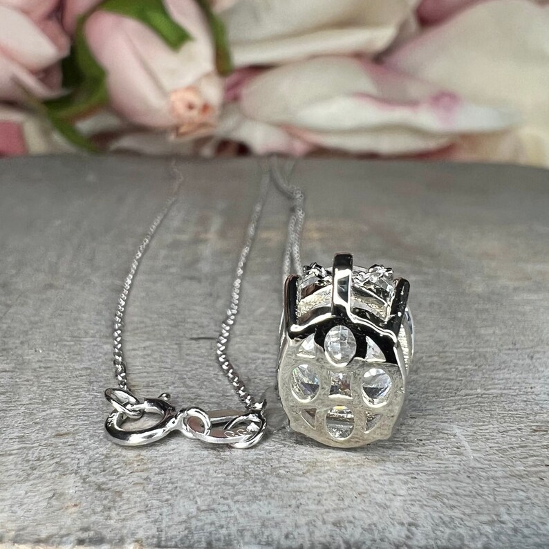 May include: A silver necklace with a pendant featuring a geometric design with clear, sparkling stones. The necklace is displayed on a gray surface with a blurred floral background.