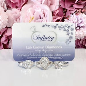 May include: Three diamond rings with different settings are displayed in front of a business card. The card has the text "Infinity Jewelers" and "Lab Grown Diamonds". The background features pink and purple flowers.