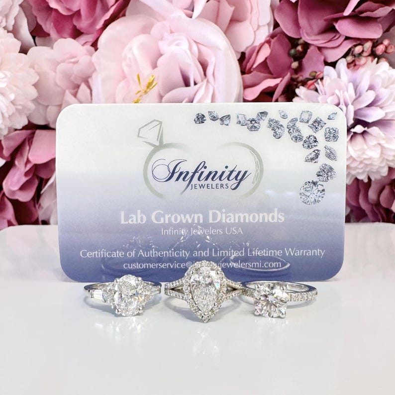 May include: Three diamond rings with different settings are displayed in front of a business card. The card has the text "Infinity Jewelers" and "Lab Grown Diamonds". The background features pink and purple flowers.