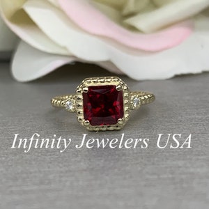 May include: A gold ring with a square red gemstone surrounded by smaller diamonds. The ring has a textured band and is set in a square halo.