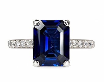 May include: A silver-toned ring featuring a large, rectangular, emerald-cut deep blue gemstone held by four prongs. The band is crafted from a metallic material and is accented with a row of small, round, clear white stones along each side.