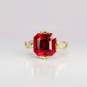 May include: A gold ring with a large, red, square-shaped gemstone surrounded by smaller, clear gemstones.