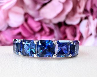 Op de afbeelding: Een zilveren ring met vijf vierkant geslepen, diepblauwe edelstenen. De edelstenen zijn dicht op elkaar geplaatst, waardoor een doorlopende band van kleur ontstaat. De ring wordt gepresenteerd tegen een zachte, vage achtergrond van roze en paarse bloemen.