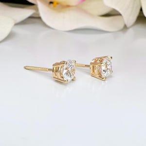 May include: A pair of gold stud earrings with clear, round-cut gemstones. The earrings are set in a four-prong setting, and the posts have a threaded design. The earrings are displayed on a white surface.
