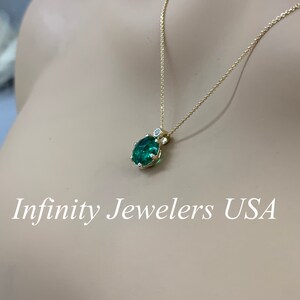 May include: A gold chain necklace with a green gemstone pendant. The pendant is surrounded by smaller diamonds. The necklace is on a mannequin's neck. Infinity Jewelers USA is written below the image.