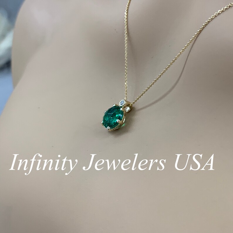 May include: A gold chain necklace with a green gemstone pendant. The pendant is surrounded by smaller diamonds. The necklace is on a mannequin's neck. Infinity Jewelers USA is written below the image.