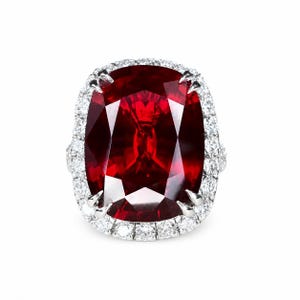 May include: A large, rectangular-cut ruby gemstone ring. The deep red ruby is surrounded by a halo of small, sparkling diamonds set in a white metal band. The ring's design features claw-like prongs securing the ruby.