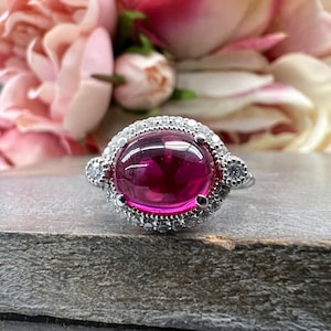 May include: A silver ring featuring a large, oval, cabochon-cut ruby gemstone. The ruby is surrounded by a halo of small, clear, round-cut diamonds. The ring is set on a gray surface, with pink flowers in the background.
