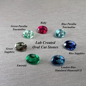May include: An assortment of oval-cut gemstones in various colors, including ruby, emerald, and sapphire. The image displays the gemstones against a gray background, with labels identifying each stone type. Text reads "Lab Created Oval Cut Stones."