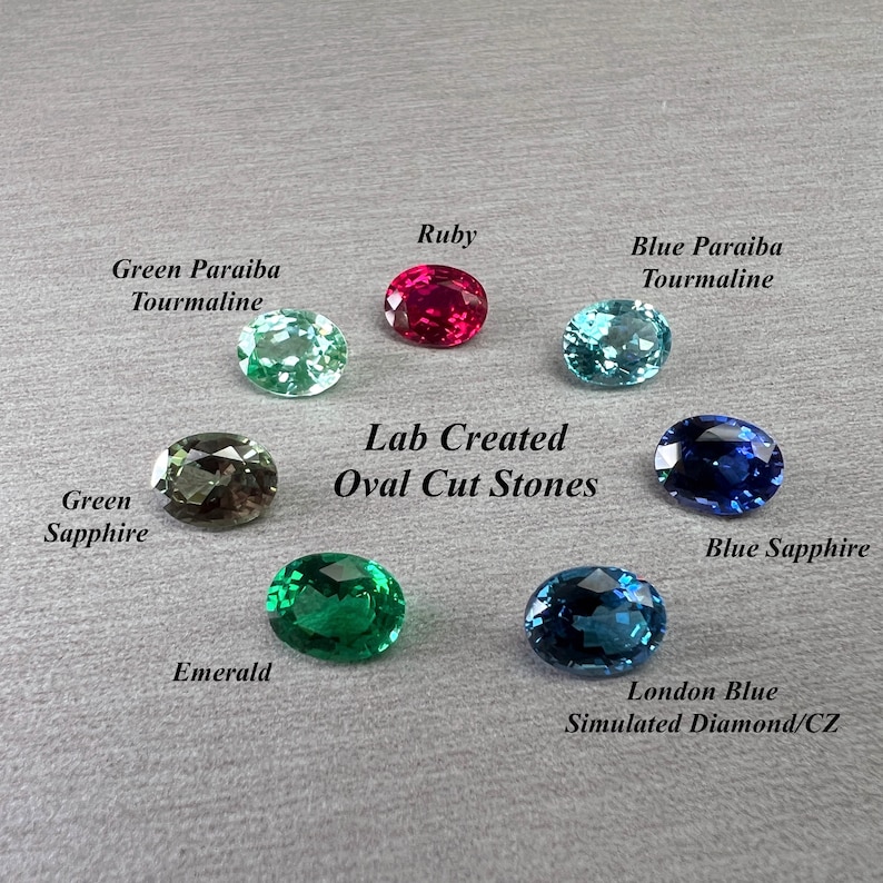 May include: An assortment of oval-cut gemstones in various colors, including ruby, emerald, and sapphire. The image displays the gemstones against a gray background, with labels identifying each stone type. Text reads "Lab Created Oval Cut Stones."