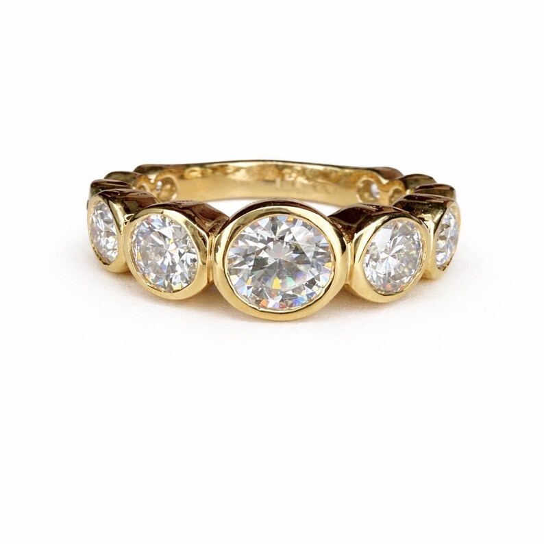 Puede incluir: Un anillo de banda dorado con m&uacute;ltiples piedras preciosas redondas y transparentes. Las piedras est&aacute;n engastadas en biseles individuales, creando un dise&ntilde;o continuo y brillante. El anillo se muestra sobre un fondo blanco, resaltando su brillo.