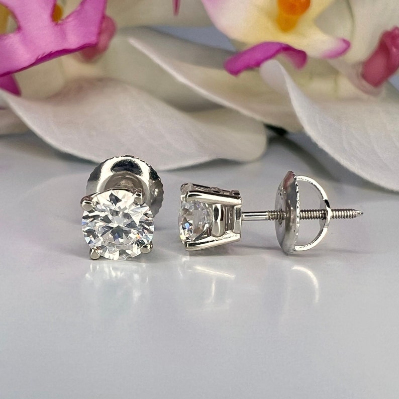 May include: Two diamond stud earrings with a bright, clear appearance. The round-cut diamonds are set in a silver metal setting. The earrings are displayed on a white surface with a blurred floral background.