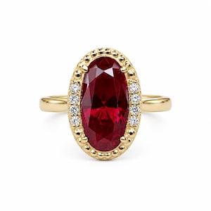 May include: A yellow gold ring featuring a large, oval-cut deep red gemstone at the center. The stone is surrounded by a beaded gold border accented with small, round, clear white diamonds set along the sides of the oval frame.