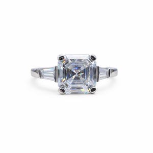 May include: A silver engagement ring featuring a large, square-cut, clear gemstone as the centerpiece. Two smaller, rectangular gemstones flank the main stone. The ring has a classic, elegant design, perfect for a special occasion.