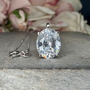 May include: A silver necklace with a large, oval-cut, clear gemstone pendant. The pendant is set in a silver bezel and has a brilliant, faceted surface. The chain is delicate and silver, with a lobster clasp.