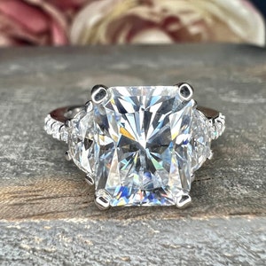 May include: A silver ring with a large, square-shaped, clear diamond in the center. Two smaller, triangular, clear diamonds are set on either side of the center diamond.