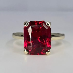 May include: A gold ring with a large, deep red, emerald-cut gemstone.