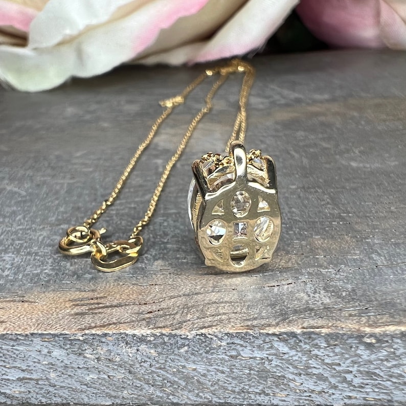 May include: A gold-colored pendant necklace with a detailed, openwork design. The pendant features multiple clear, sparkling stones set within a decorative frame. The chain is delicate and also gold-toned, creating an elegant jewelry piece.