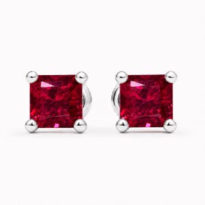 May include: A pair of stud earrings featuring square-cut, deep red gemstones set in polished silver-toned metal. Each stone is secured by four rounded prongs at the corners, creating a classic, symmetrical design with a vibrant, faceted appearance.