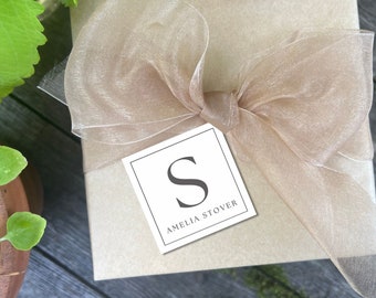 May include: A brown gift box with a beige sheer ribbon tied in a bow. A white gift tag with a black border is attached to the ribbon. The tag has a black letter "S" and the text "Amelia Stover" printed on it.