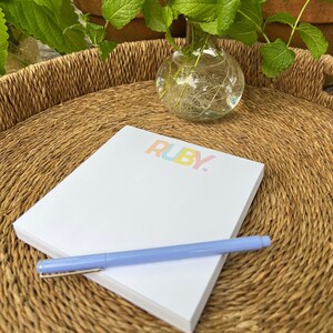 May include: A white notepad with the word "RUBY" printed in rainbow colors on the top. A blue pen is lying on top of the notepad. The notepad is sitting on a woven wicker tray.