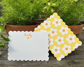 May include: Two white note cards with scalloped edges. One card has the name "Ruby" written in orange with a small yellow flower. The other card has a yellow background with white flowers with orange centers.