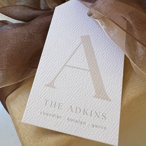 May include: A white gift tag with a textured surface. The tag features a large letter "A" in a light brown color. The text "THE ADKINS" is printed in a light brown color below the letter. The names "chandler", "katelyn", and "gotlin" are printed in a smaller font below the text.